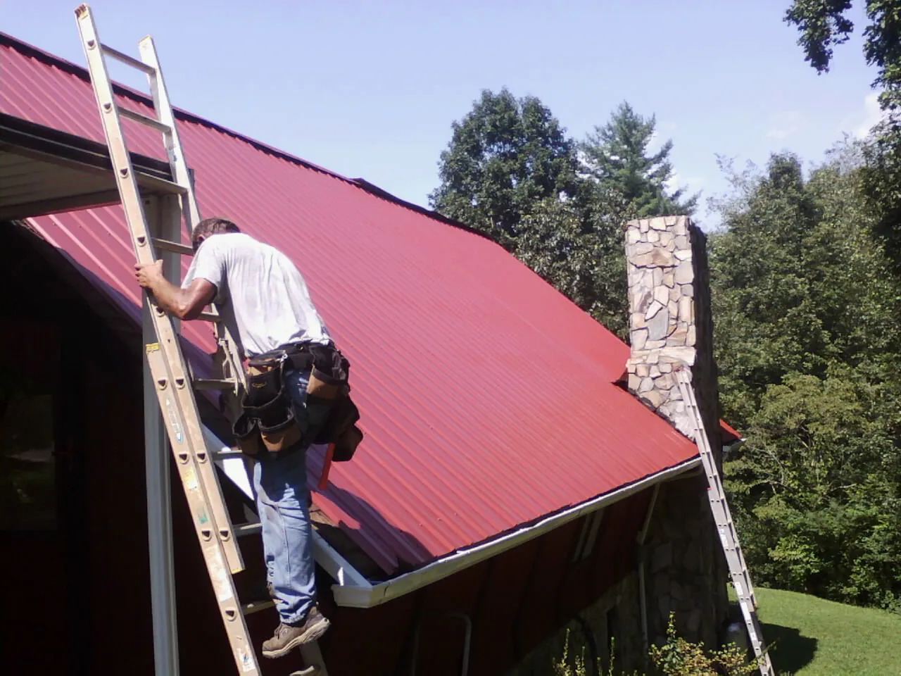 Roofer on a ladder beside a red metal roof and stone chimney — Soffit & Fascia Repair by Roofing Brothers in Navasota